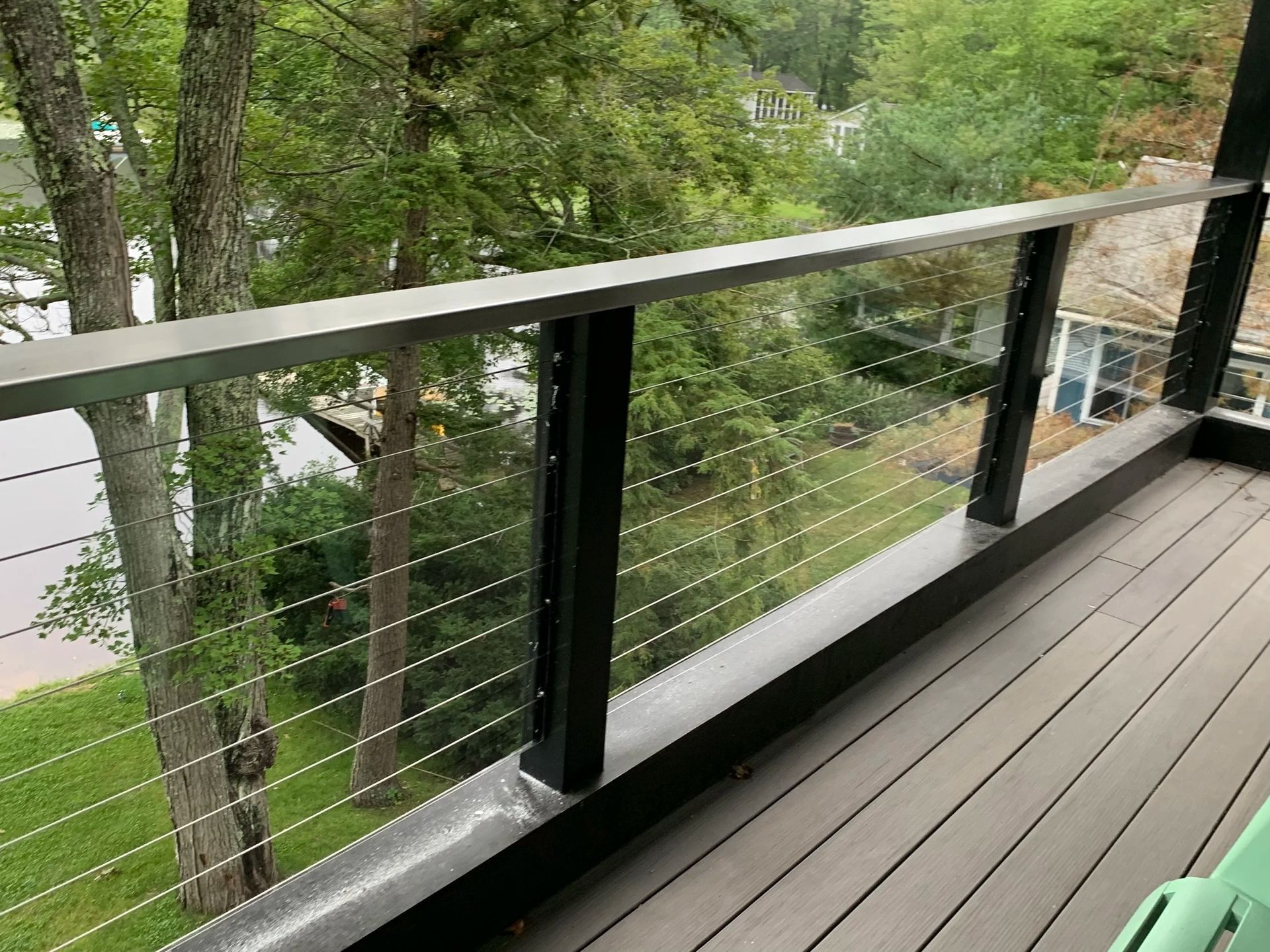 Deck railing with horizontal metal cables, black posts, and a gray top rail. Green trees and water in the background.