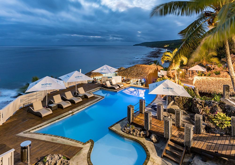 Resort infinity pool at sunset overlooking the ocean, with lounge chairs, umbrellas, and palm trees