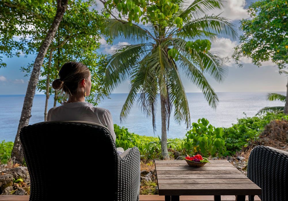 Person seated on a patio overlooking palm trees and the ocean, with a small table and flowers nearby
