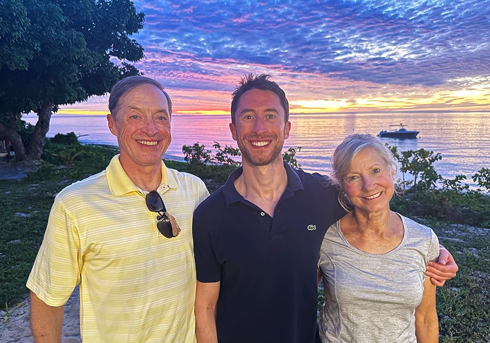 Three smiling people standing outdoors at sunset by the water, with a colorful sky and a boat in the distance