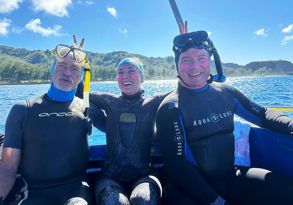 Three snorkelers in wetsuits smiling on a boat with blue ocean and sunny coastline behind them