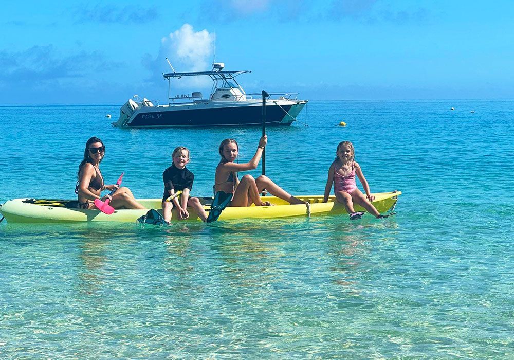 Four people on a yellow kayak in clear turquoise water, with a boat in the background.