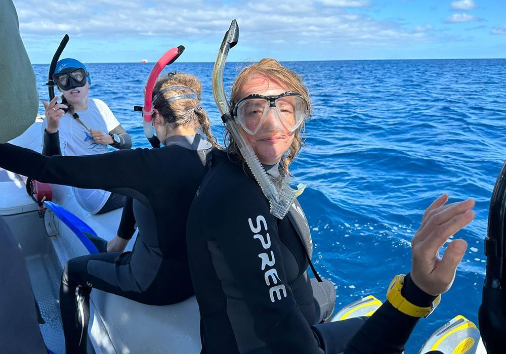 Three snorkelers on a boat in blue ocean, one smiling and waving in a wetsuit and mask.