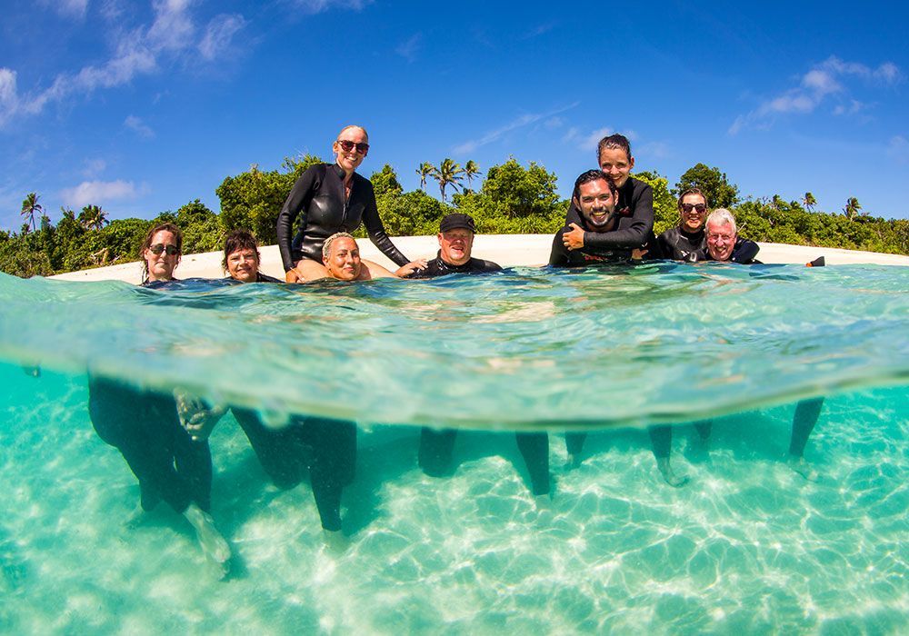 People standing in shallow turquoise water at a tropical beach, smiling and leaning on a floating edge