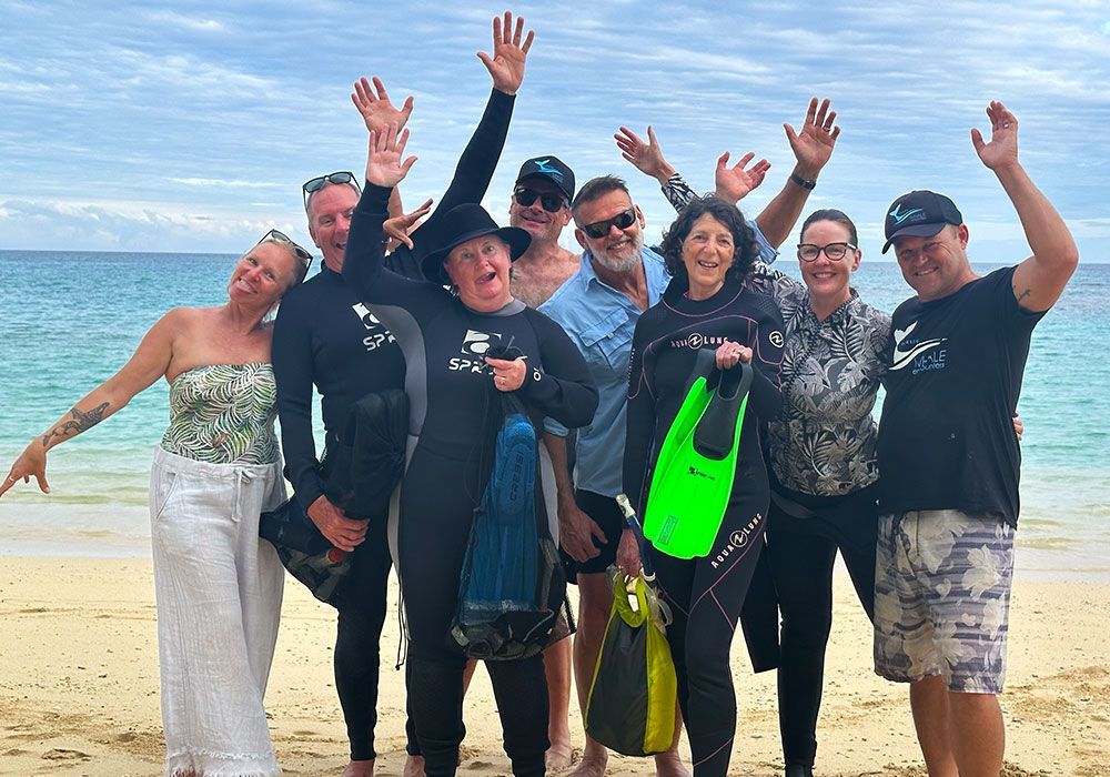 Group of people in wetsuits and swimwear waving on a sunny beach with turquoise water behind them
