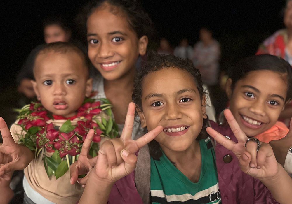 Four smiling children posing close together, two making peace signs, in a dimly lit setting.