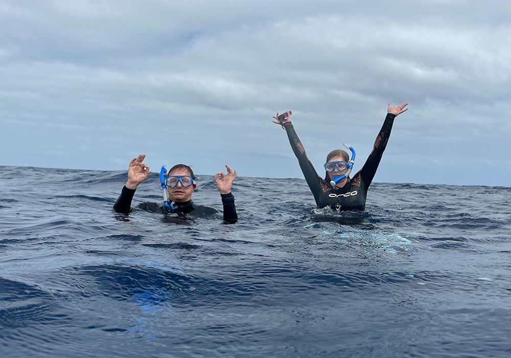 Two snorkelers in wetsuits and masks bob in open water, raising their arms under a cloudy sky.