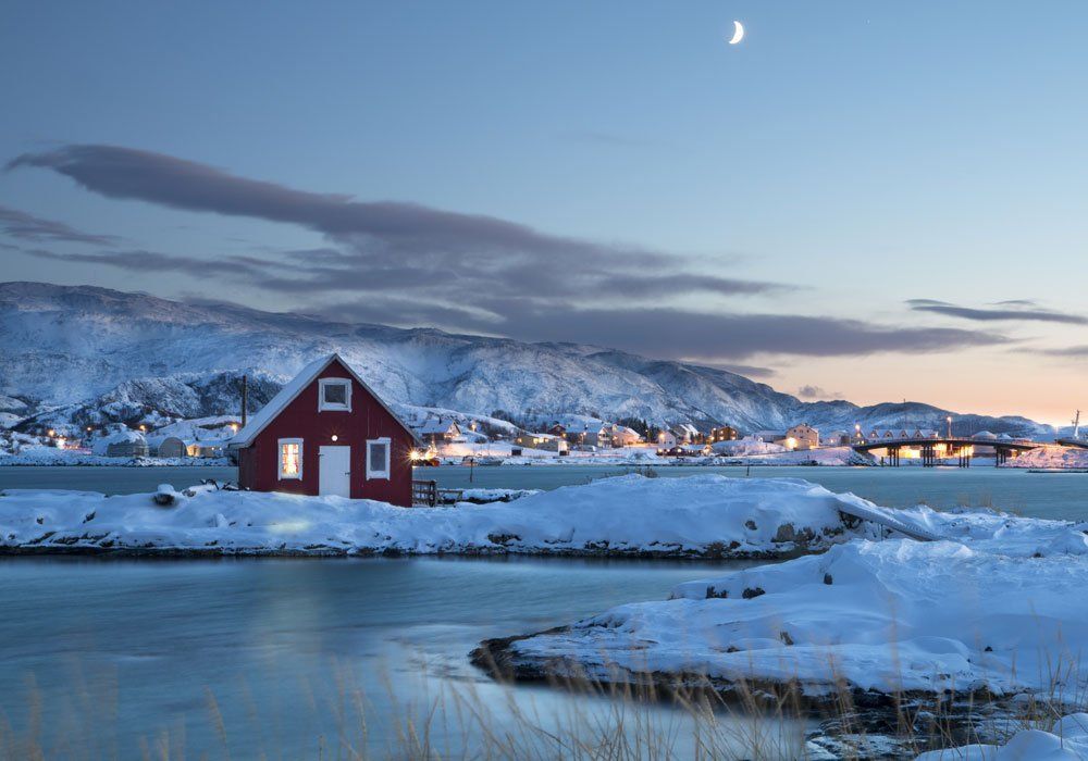 harbour A red house is sitting on a snow covered island next to a body of water.