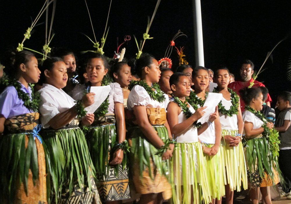 Group of performers in green grass skirts and floral leis singing on stage at night