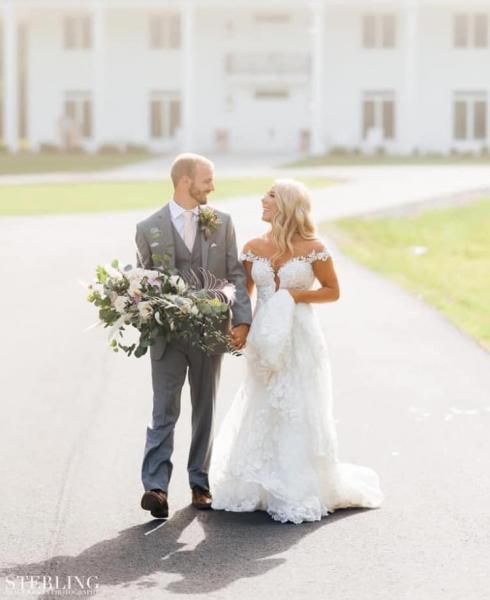 Bride and groom walking hand in hand outdoors, holding a bouquet in bright sunlight