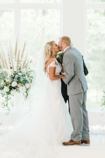 Bride and groom kissing at a bright wedding venue beside a white floral arrangement