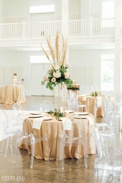 Elegant wedding reception table with beige linens, white chairs, and a tall floral centerpiece in a bright room