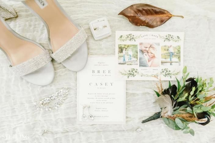 Wedding invitation flat lay with white lace shoes, envelope, floral print card, leaf, and greenery on a white background