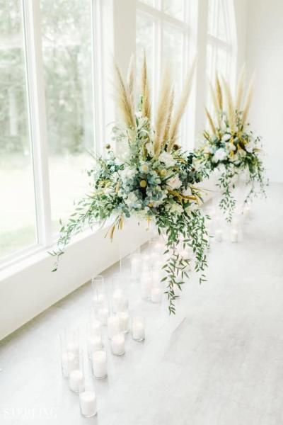 White floral arrangements with tall pampas grass beside a bright window, with candles on the floor