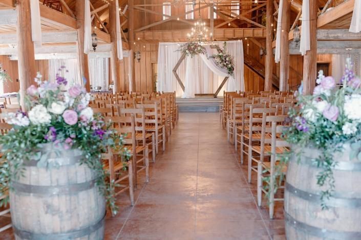 Decorated rustic wedding aisle with wooden chairs, floral arrangements, and draped altar in a barn
