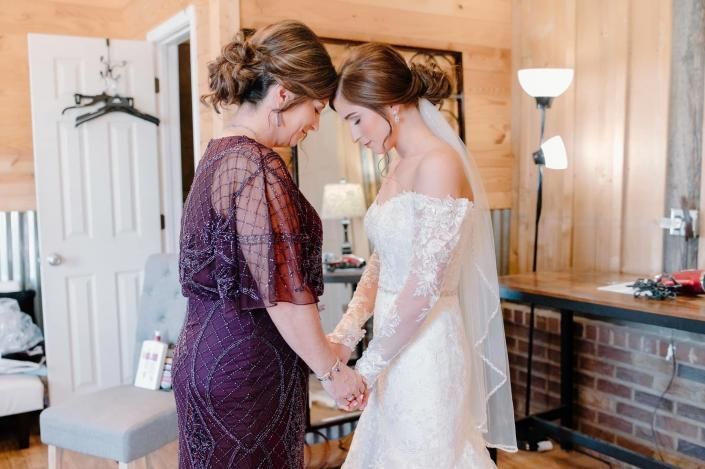Bride and woman in purple dress hold hands indoors, sharing an emotional moment near a fireplace