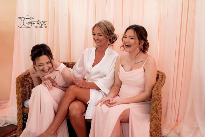 Three women in pink and white dresses seated together on a sofa, smiling in a softly draped room.