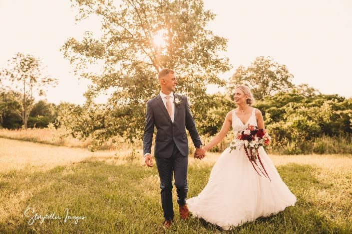 Bride and groom walking hand in hand in a sunny meadow, with a tree and warm golden light behind them.