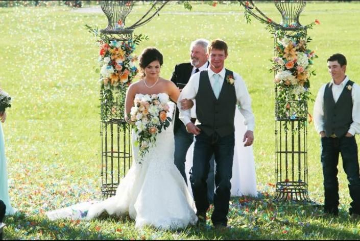 Bride and groom walking down an outdoor aisle under a floral arch, with attendants nearby