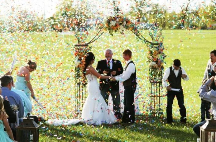 Outdoor wedding ceremony with bride and groom under a flower arch as confetti falls around them