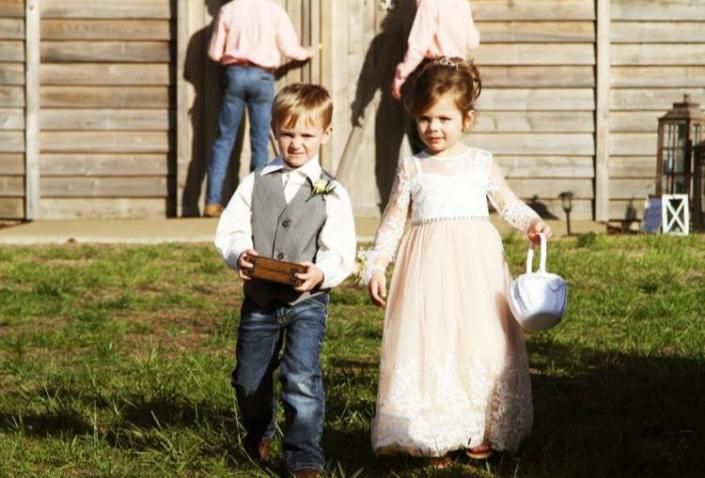 Two children walking on grass, a boy carrying a box and a girl holding a small bucket near a wooden building.