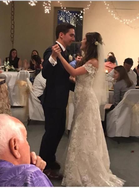 Couple slow dancing at a wedding reception, with guests seated at decorated tables nearby.
