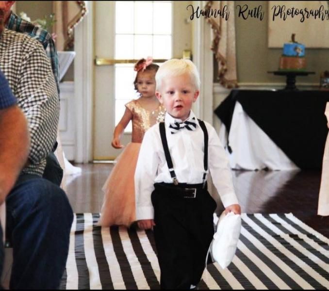 Child in black suspenders walks down an indoor striped aisle, with another child in a peach dress behind.