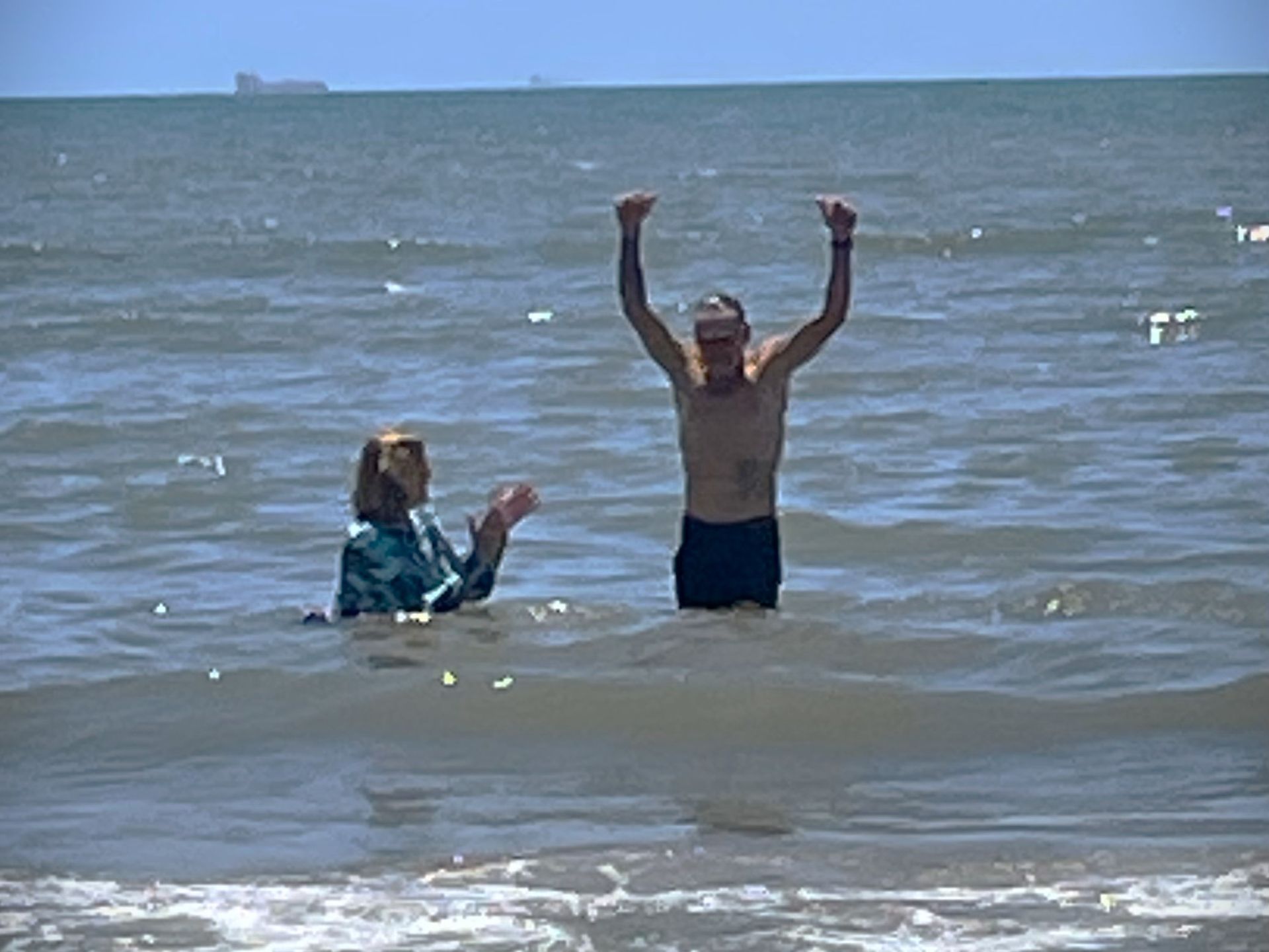 Two people in a lake, one standing with arms raised and one sitting in shallow water near the shore