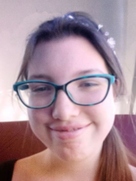 Smiling person wearing teal glasses, close-up selfie against a light background.