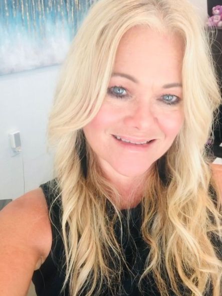 Close-up selfie of a smiling blonde woman in a black sleeveless top indoors