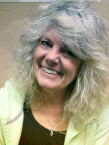 Smiling woman with curly gray hair wearing a light jacket and dark top on a plain background