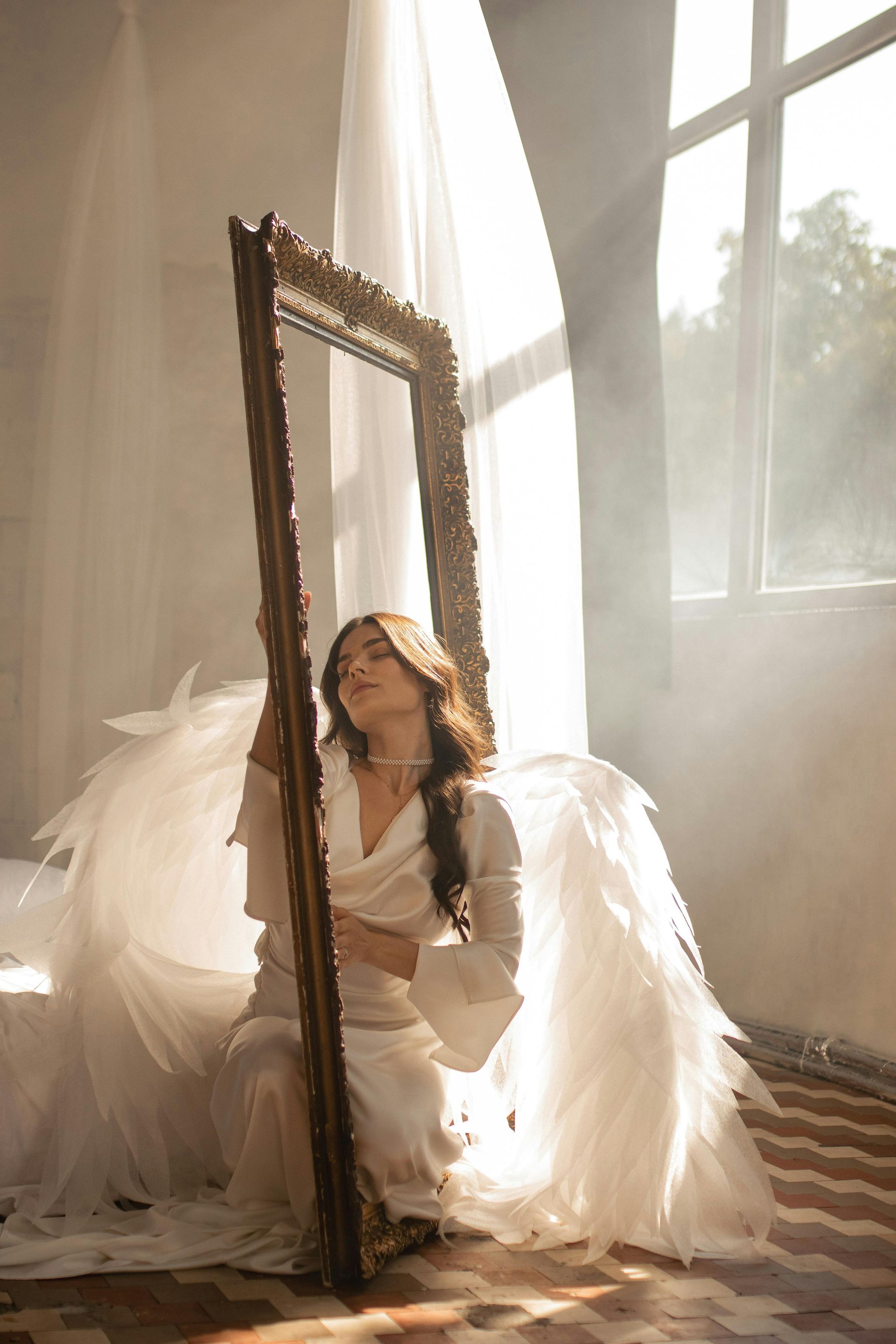 Woman with large white wings, in a white robe, holds a gold framed mirror in a sunlit room.