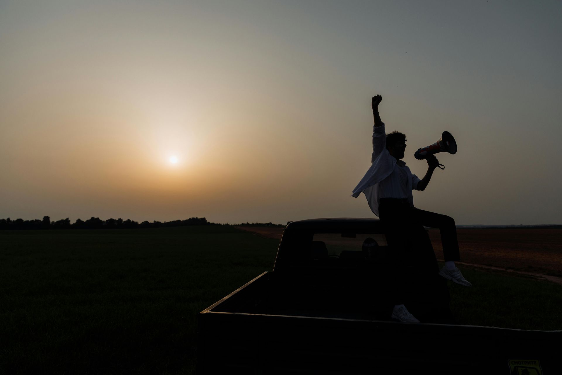 Silhouette of a person on a truck bed holding a megaphone, arm raised in a field at sunset.
