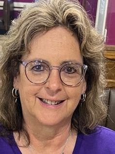Portrait of a smiling woman with glasses and hoop earrings, wearing a purple top.