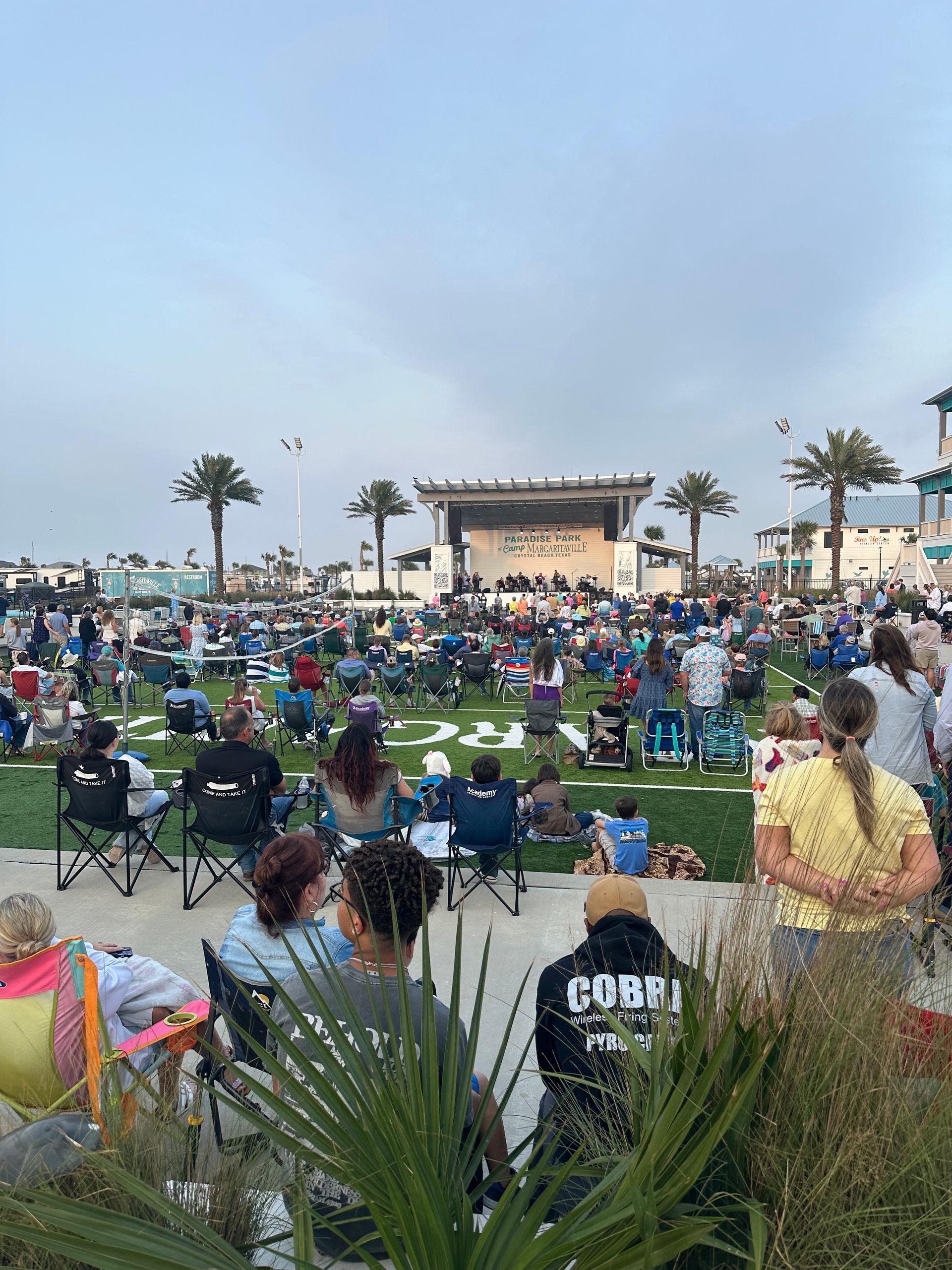Crowd seated on lawn facing an outdoor stage at dusk, with palm trees and folding chairs.