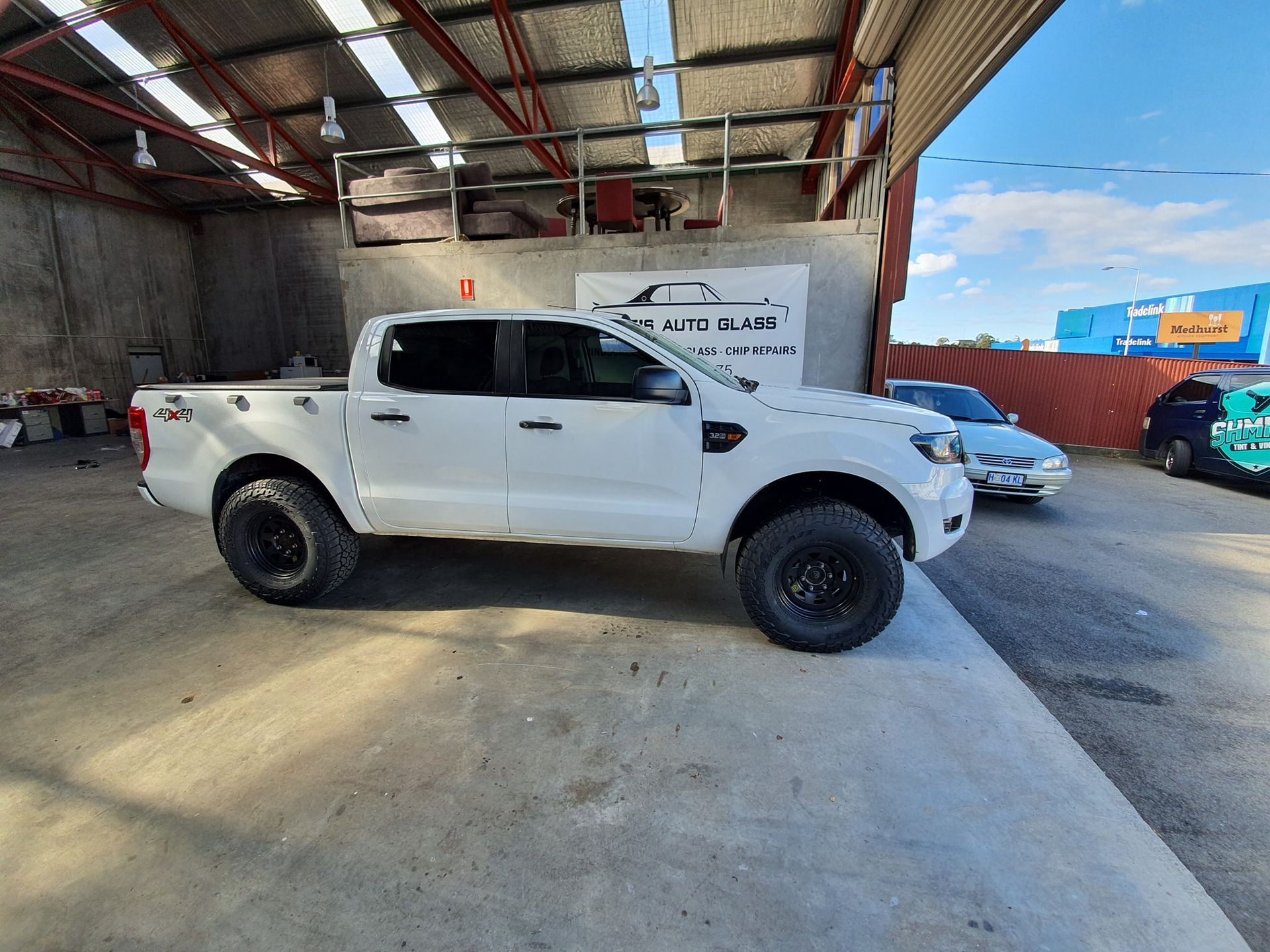 White pickup truck parked in a warehouse, with a white car in the background and blue sky visible outside.