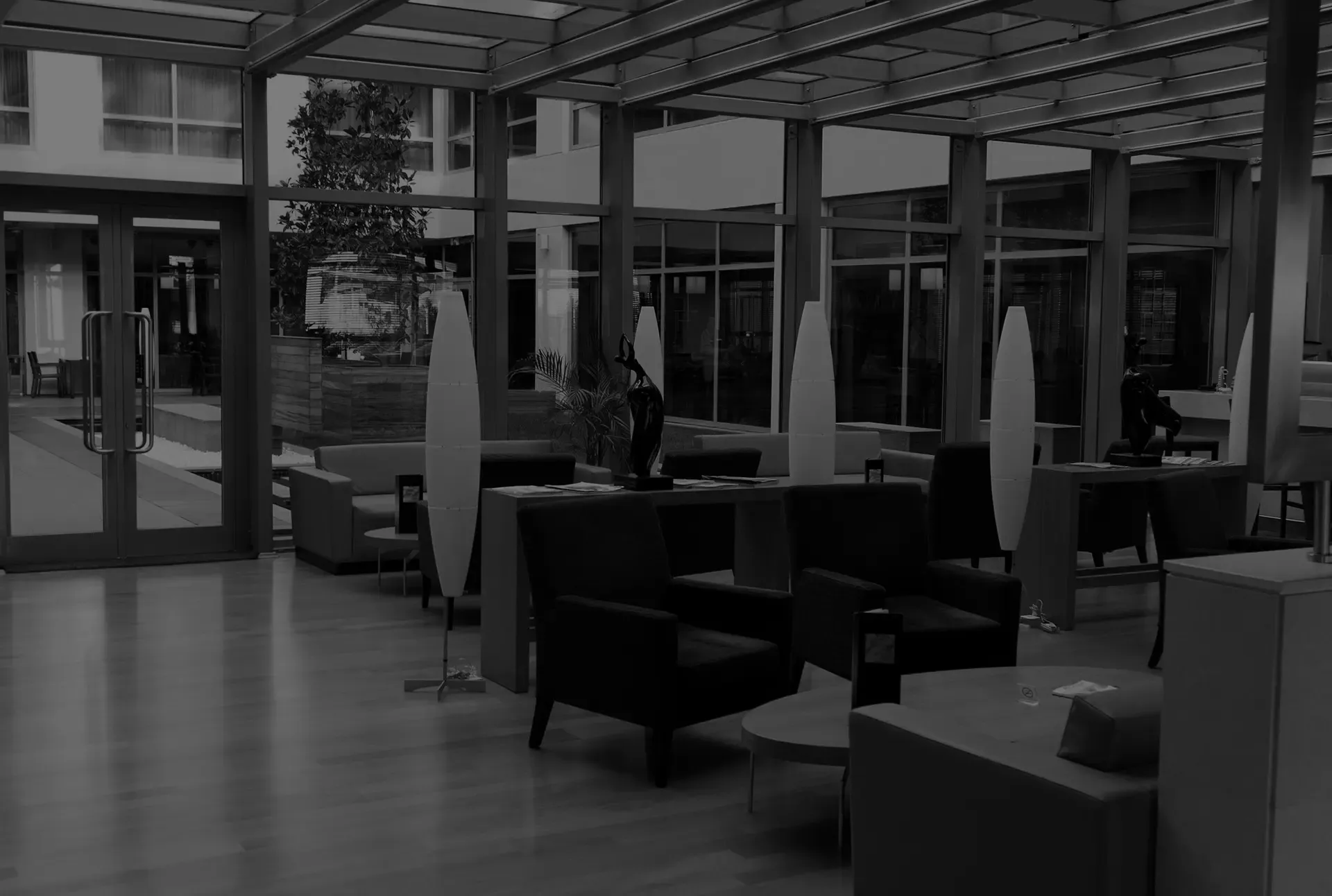 Lounge area with chairs, small tables, and tall lamps. Glass walls and doors offer a view to the outside.