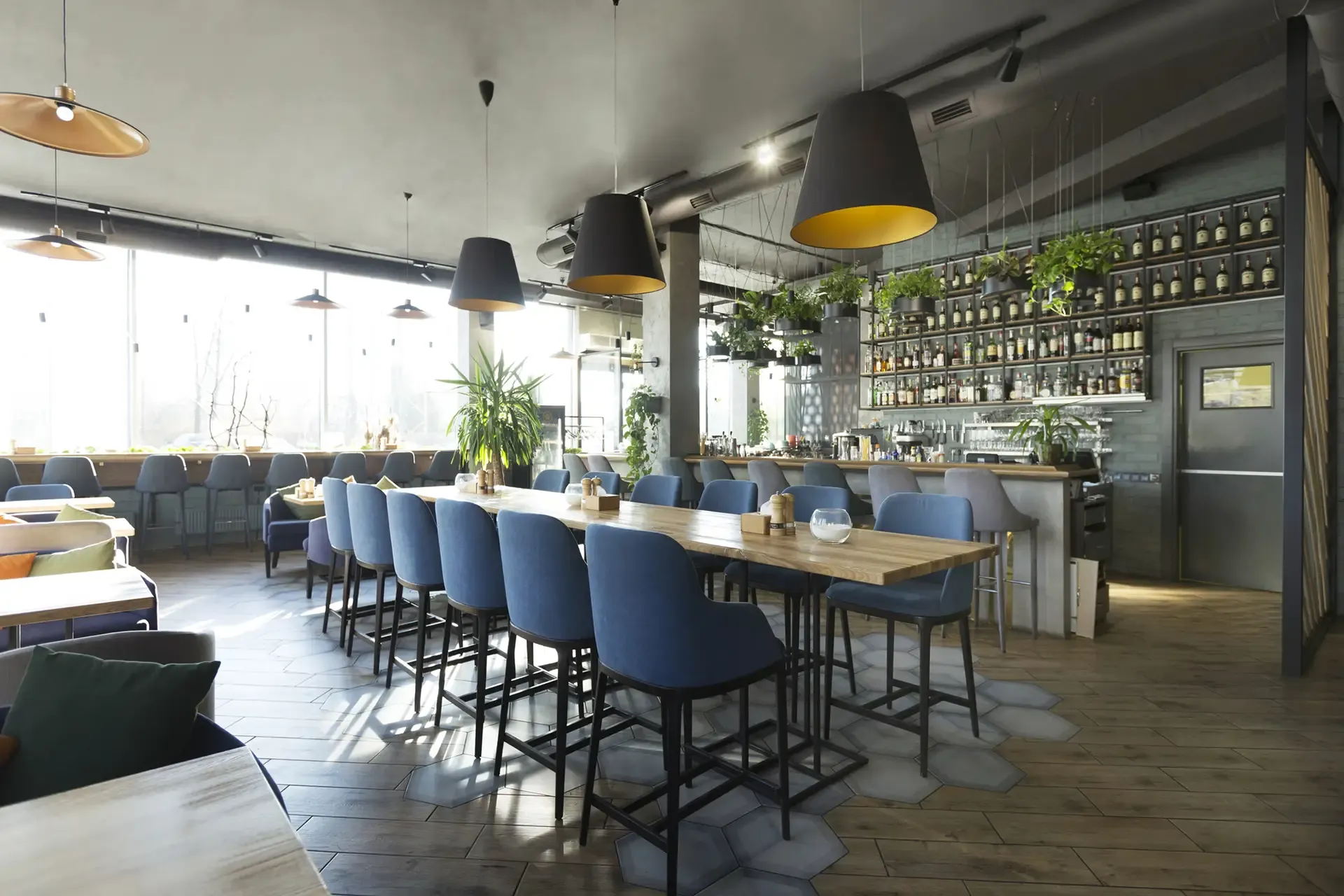 Restaurant interior with long tables, blue chairs, a bar, and hanging lamps.