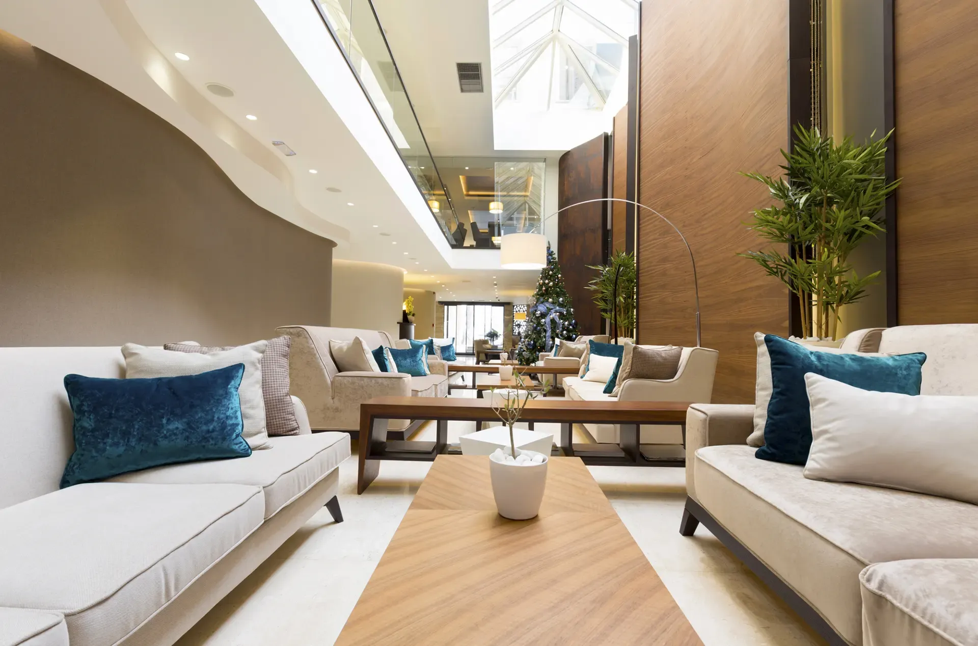 Lobby with couches, tables, and plants. Features teal pillows, wood accents, and a high ceiling with skylights.
