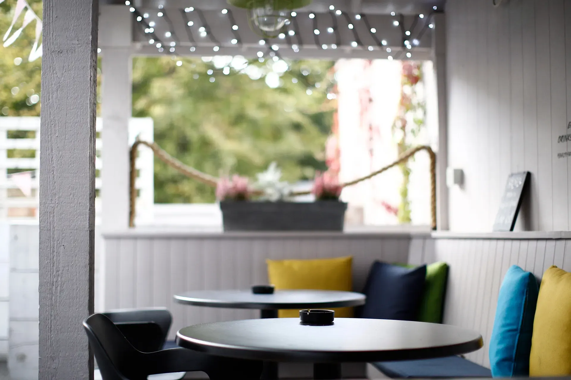 Outdoor seating area with round tables, colorful cushions, and a blurred background of greenery.