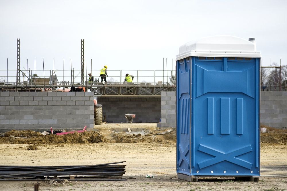 Blue portable toilet at an active construction site with workers, concrete block walls, and building materials.