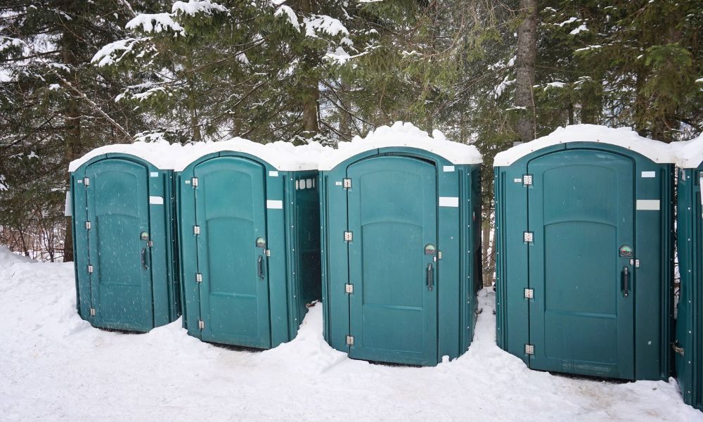 Green portable toilets surrounded by snow at a wooded outdoor site during freezing winter weather.