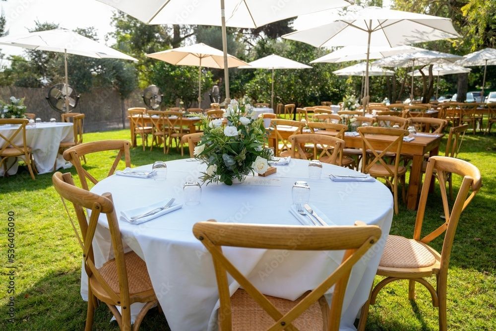 Tables and chairs are set up for a party in the grass under umbrellas.