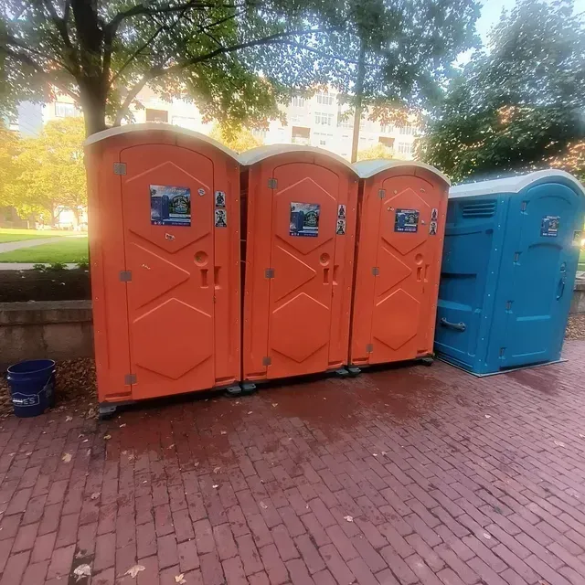 Four portable toilets set up outdoors on a brick-paved area for an event or job site.
