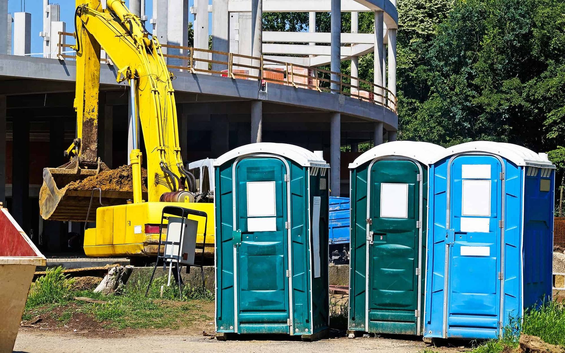 Three portable toilets positioned near an excavator and partially constructed commercial building for convenient job site access.