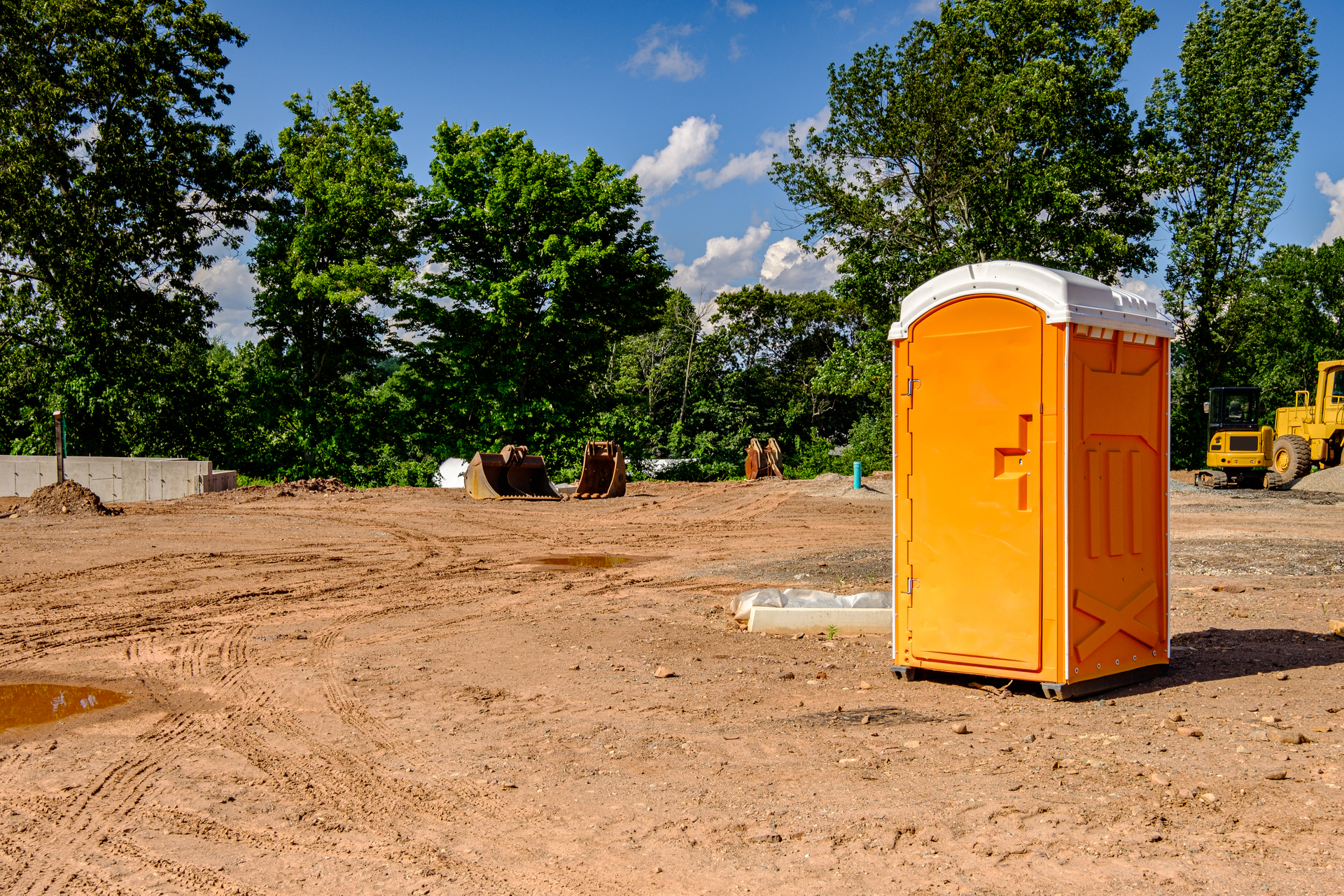 Orange portable toilet positioned on level ground at a construction site with excavation equipment i