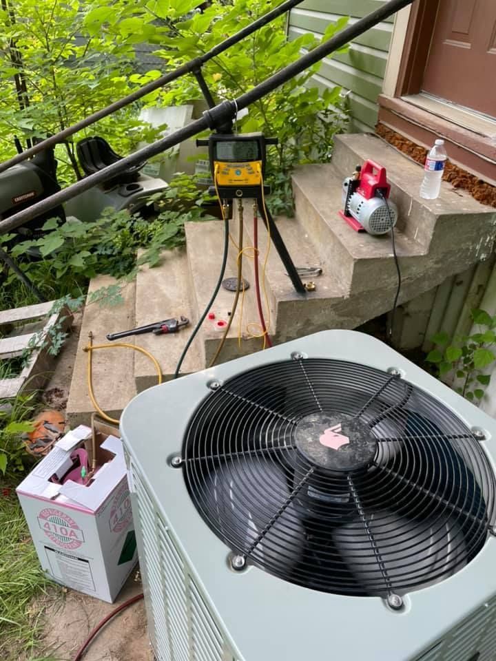 Outdoor HVAC condenser and fan with repair tools on a porch beside green plants.