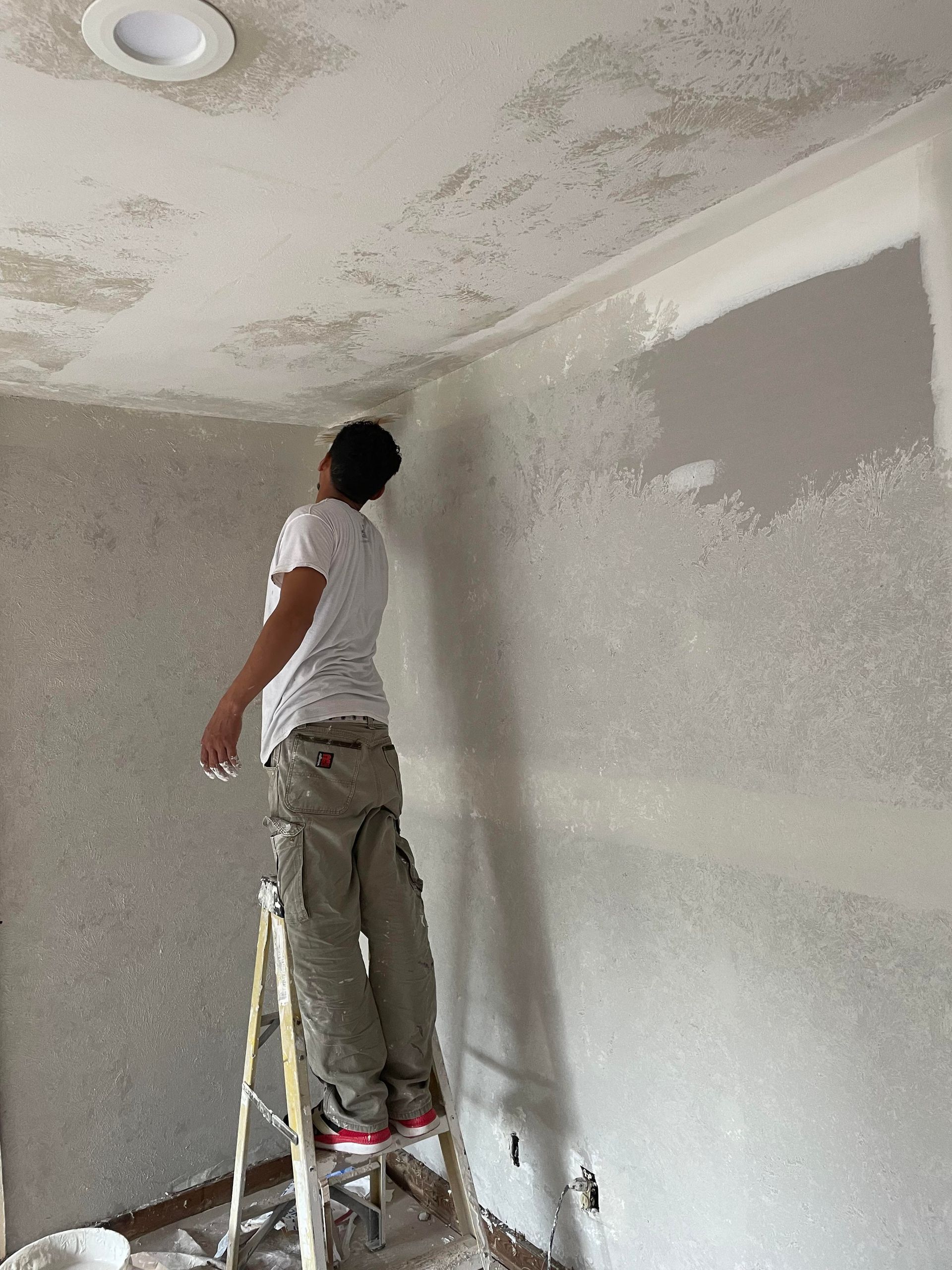 Person standing on a ladder painting a white ceiling and wall in a room