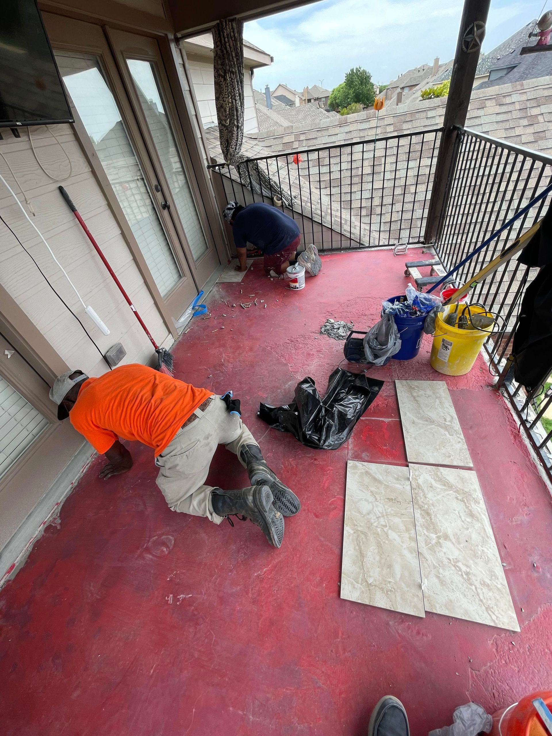 Two workers cleaning a red porch floor with tools and supplies scattered nearby