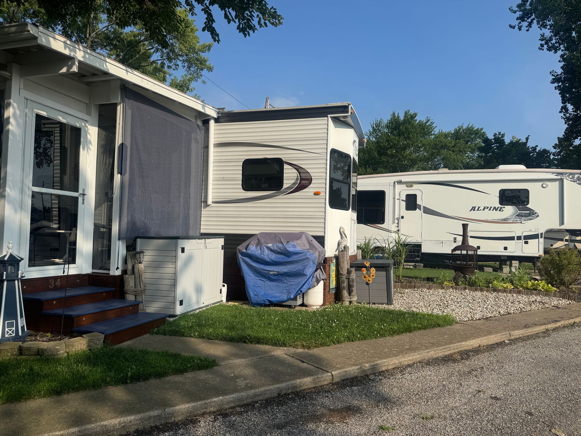 Small white RV homes on a gravel lot beside a house, with blue sky and trees in the background